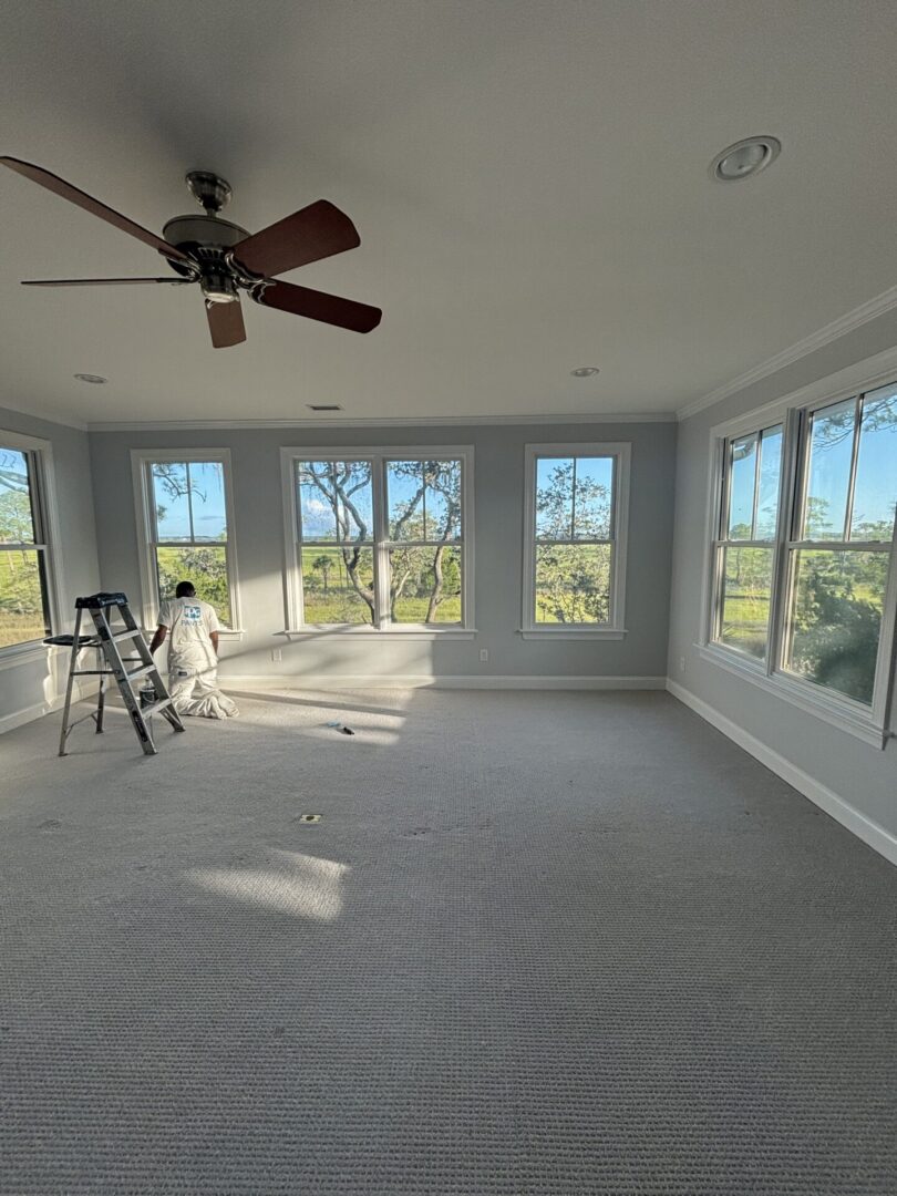 Professional interior painter finishing gray walls and white trim in a bright room with large windows and natural light.